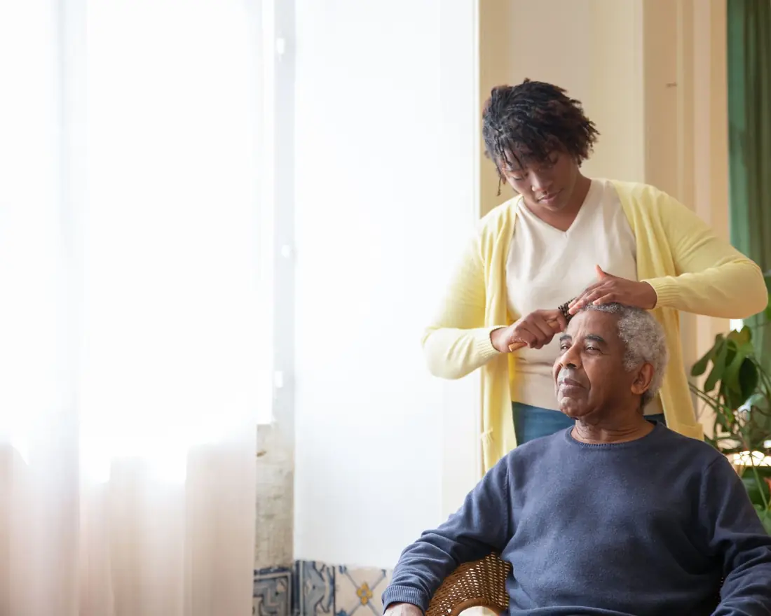 A caregiver gently combing an older man's hair by a sunlit window