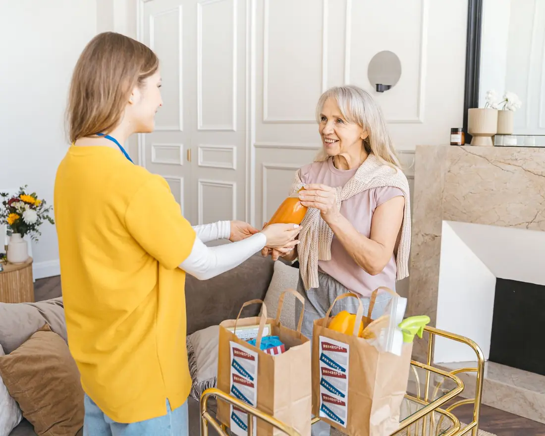A caregiver handing groceries to a smiling older woman in her living room